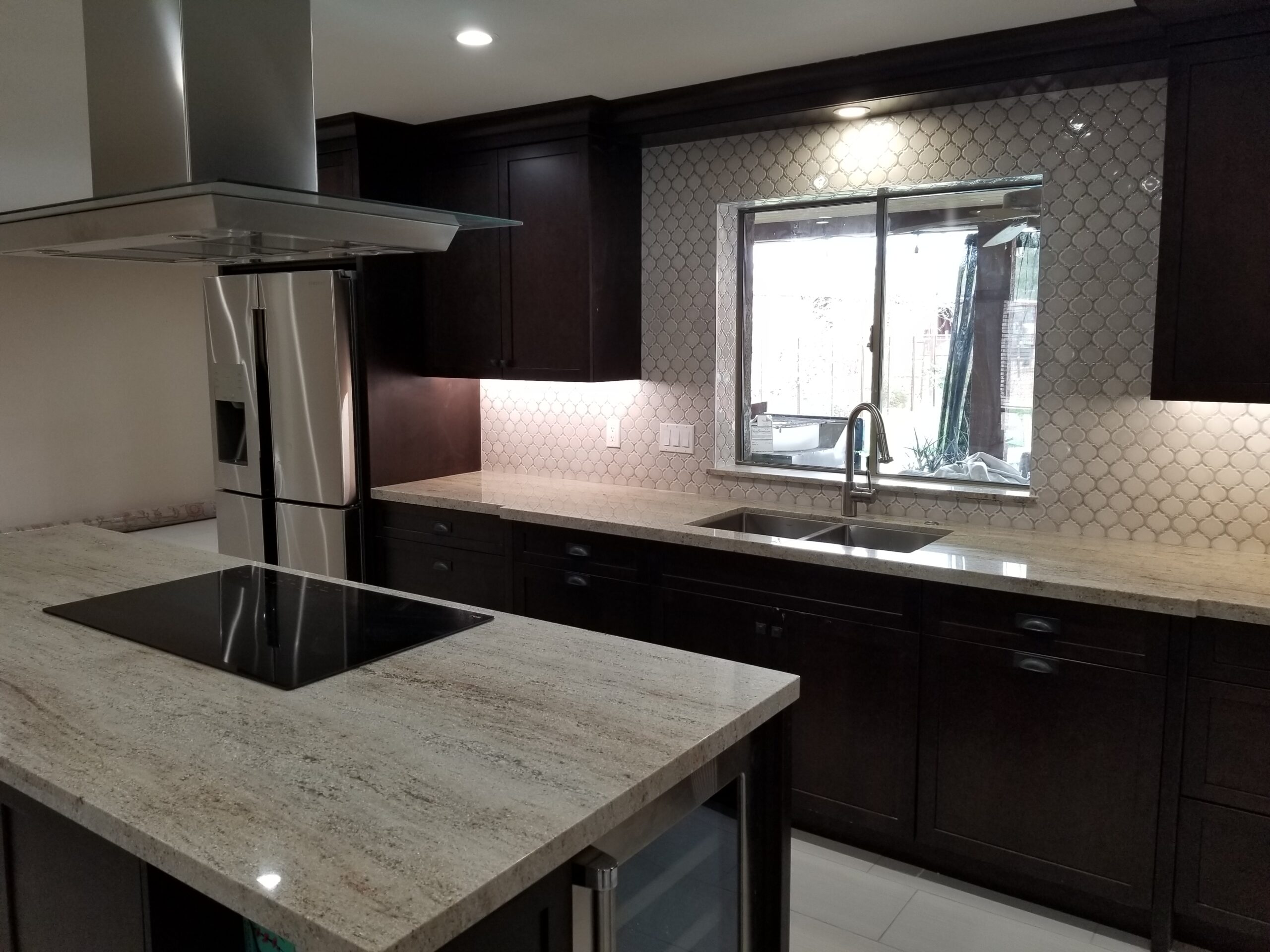 Another view of a modern kitchen in a Mesa, Arizona home with dark espresso cabinets, a light granite countertop island with a built-in cooktop, stainless steel appliances, and a patterned backsplash.