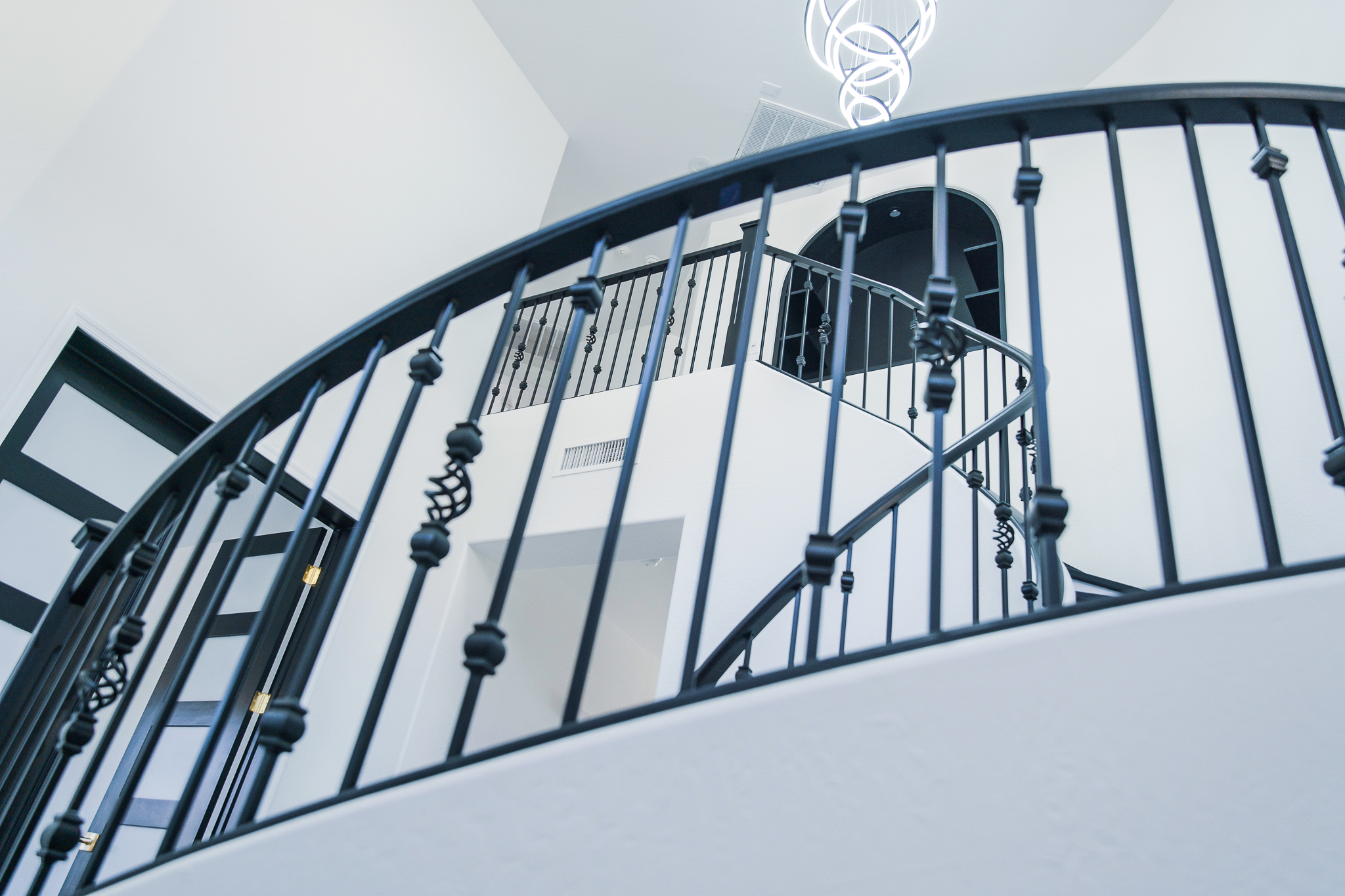 Close-up of a curved staircase with ornate black wrought iron railings in a bright Tempe entryway, with an Artemide Alphabet of Light chandelier visible above.