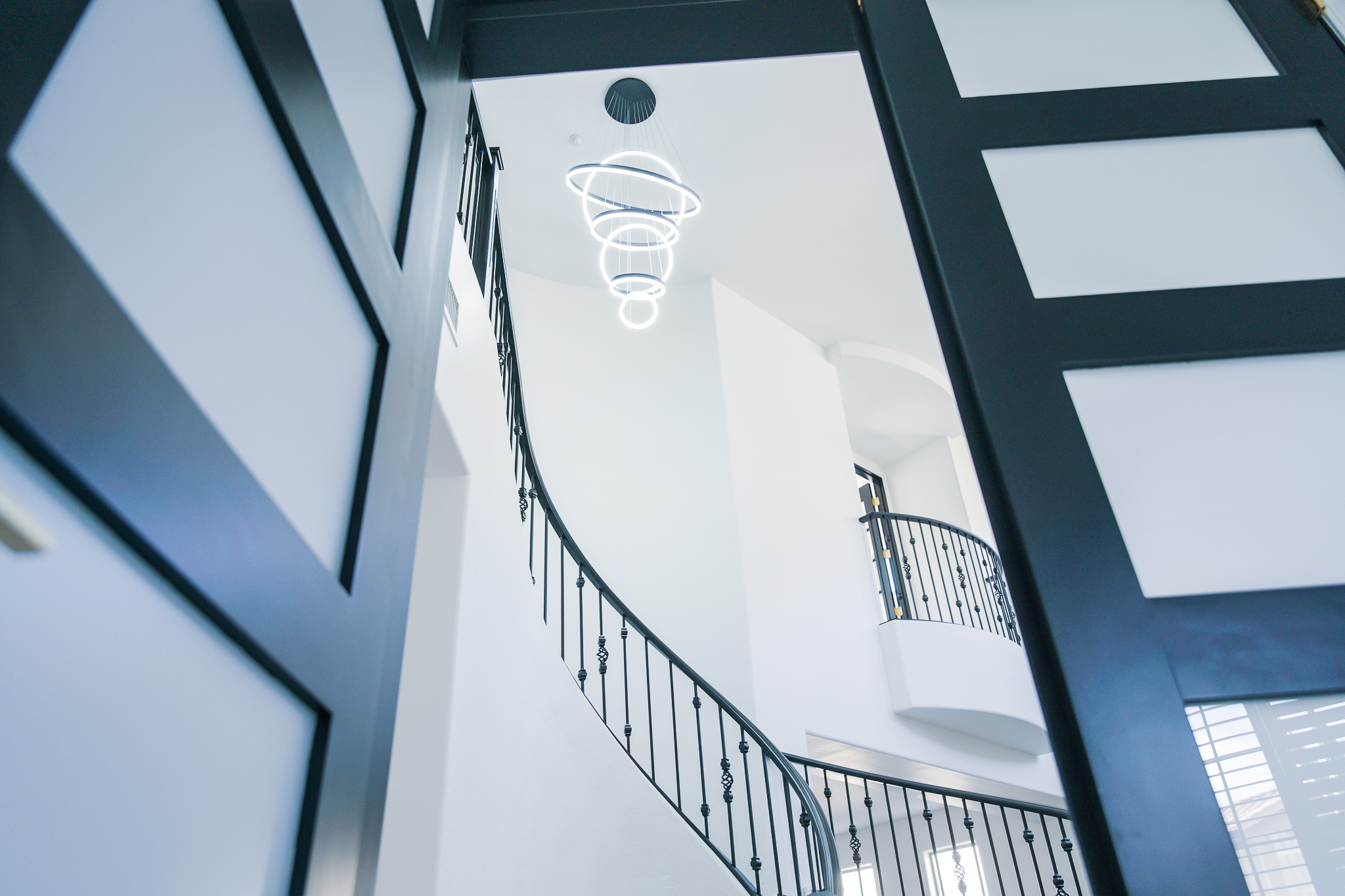 Angled view from the lower level of a Tempe home's entryway, looking up at a modern Artemide Alphabet of Light chandelier and a curved staircase with black railings.