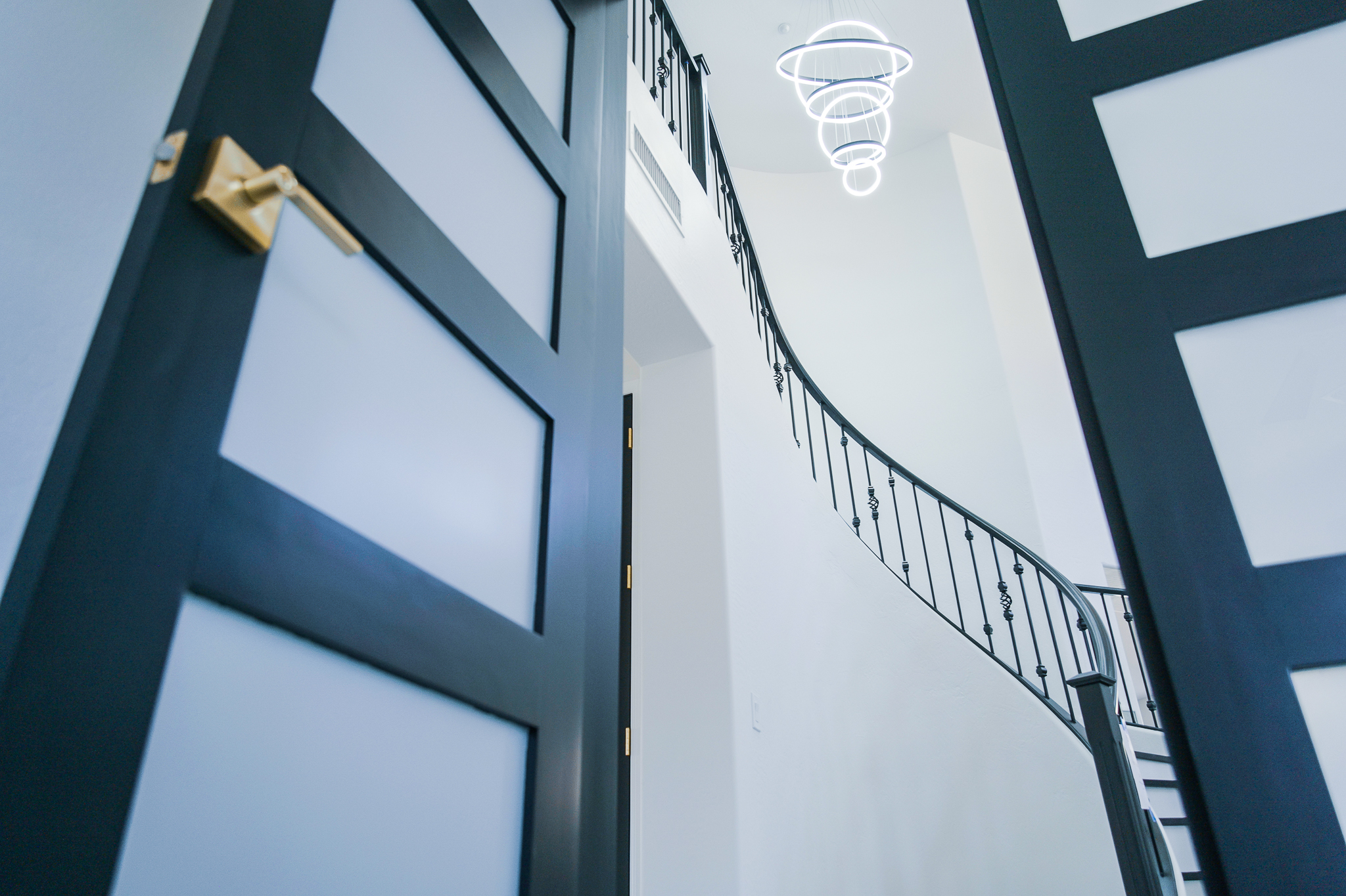 Angled view into a bright Tempe entryway, showcasing a modern LED chandelier, a curved staircase with black railings, and a partially open black and white patterned door.
