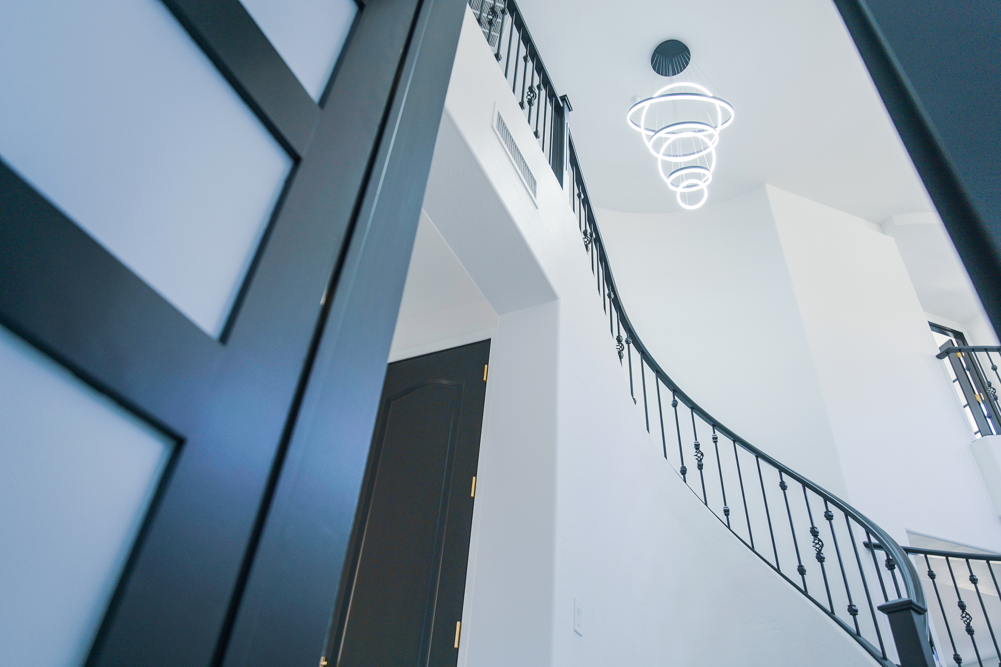 View from the lower level of a Tempe home's entryway, looking up at a modern LED chandelier and a curved staircase with black railings.