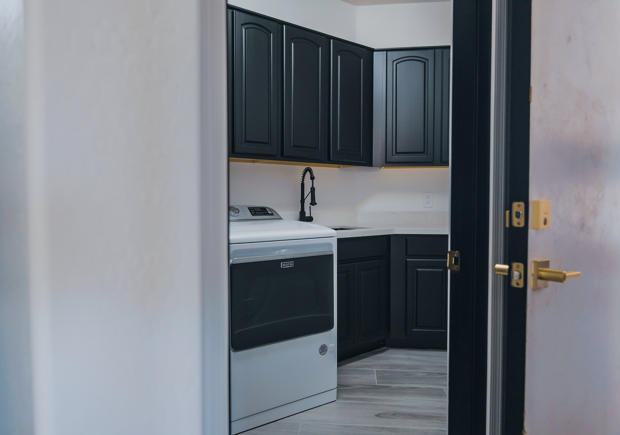 Laundry space with a white Maytag dryer, sleek black cabinets, light countertops, and a black Kraus Bolden faucet.