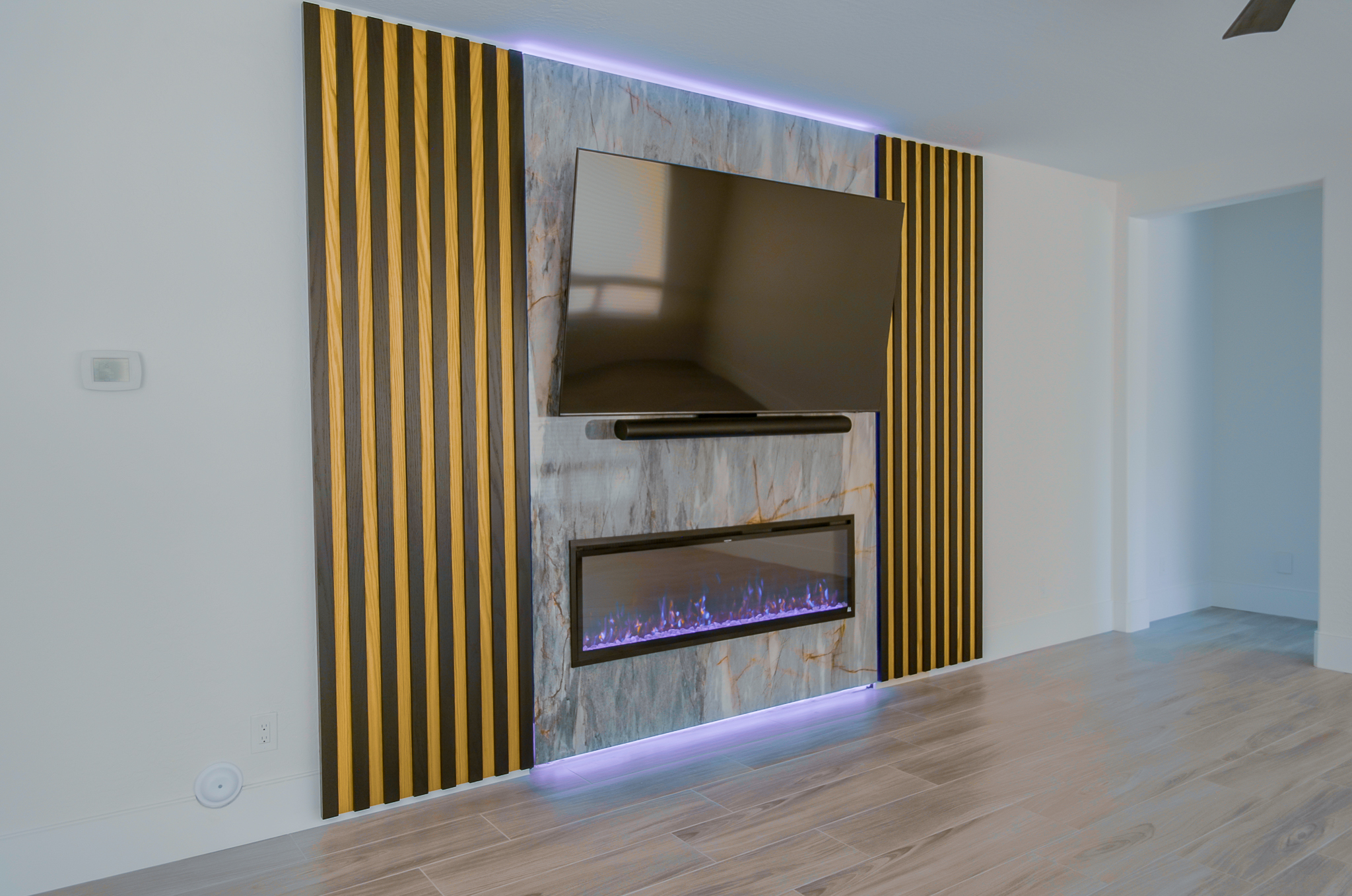 Living space with a stone and wood slat accent wall, housing a TV and electric fireplace with purple flames.