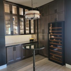 Stylish Home Bar and Wine Nook with Custom Cabinetry A modern home bar or butler's pantry area featuring dark cabinetry, a countertop with glass-front display cabinets above, a freestanding wine refrigerator, and a small round bistro table with a dark base and top, illuminated by an overhead drum pendant light.