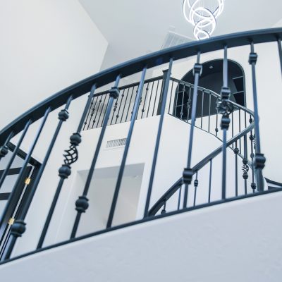 Close-up of a curved staircase with ornate black wrought iron railings in a bright Tempe entryway, with an Artemide Alphabet of Light chandelier visible above.