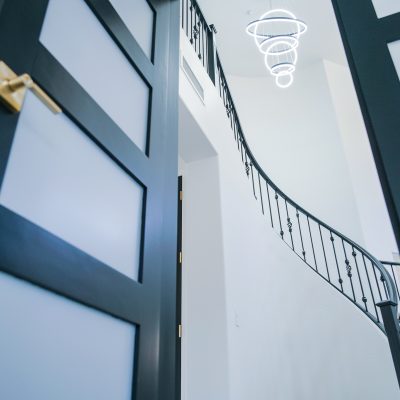 Angled view into a bright Tempe entryway, showcasing a modern LED chandelier, a curved staircase with black railings, and a partially open black and white patterned door.