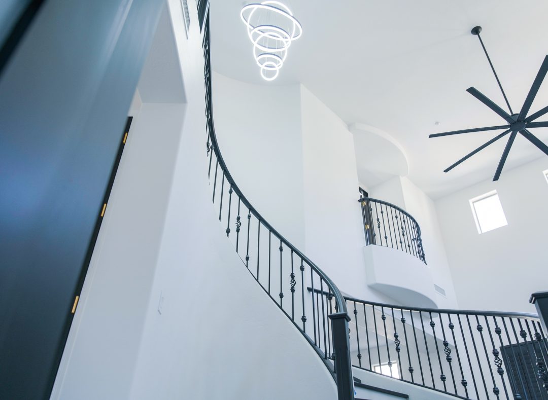Grand Foyer with Spiral Staircase and Modern Lighting A dramatic, low-angle view of a luxurious, multi-story foyer featuring a prominent curved black wrought iron staircase, a contemporary white multi-ring chandelier suspended from a high ceiling, and a large black ceiling fan in the distance.