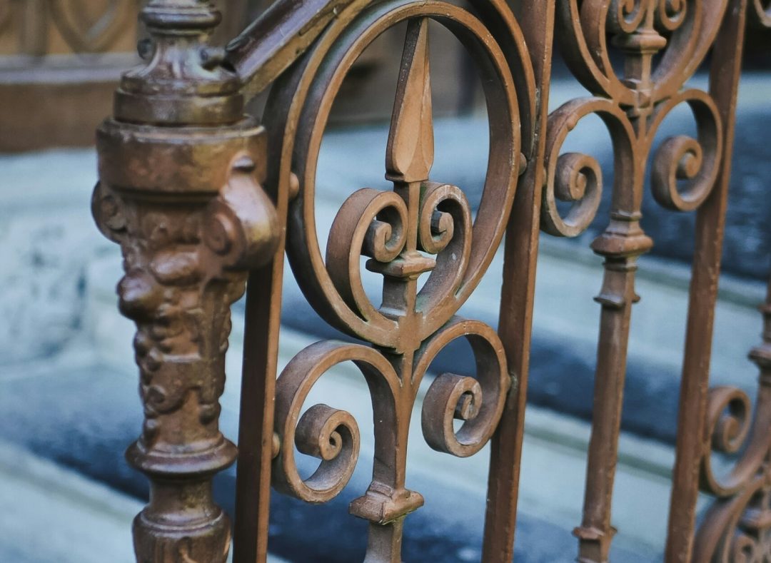 Ornate Wrought Iron Railing in Tempe, AZ Close-up of an ornate, rusty brown wrought iron railing with decorative scrolls and finials, set against stone steps, possibly in a historic building in Tempe, AZ.